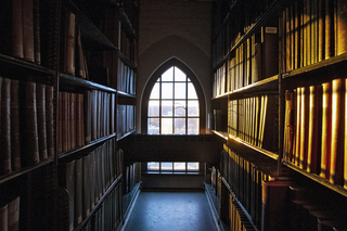 Dark aisle between shelves of books in a library looking towards an arched window