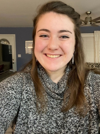Alexis, a pale white woman with dark, long brown hair and blue eyes, smiles at the camera. She is wearing dangly silver earrings and a black and white sweater with a turtleneck.