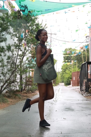 A tall, slender dark skinned Black woman wearing a black tank top and olive green pants carrying an olive green Strand Books tote bag, is facing the camera sideways with a small smile, looking as if she is heading somewhere. She has gold hoops, gold rimmed glasses on, and a gold bead in her hair, which is pulled back into a low knot.