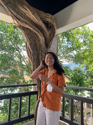 K, an East Asian person with long black hair, stands in front of trees and distant mountains in Taiwan, with their right hand near their face as they smile at the camera. They are wearing an orange shirt and cream colored pants.