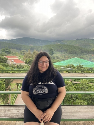 Lusangelis is sitting on a bench smiling warmly as she looks at the camera on a cloudy day with the background showing the mountains of Da Lat, Vietnam.