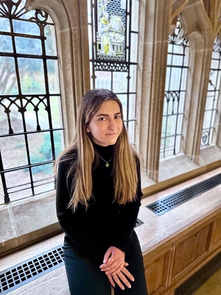 Sara, wearing a black turtleneck and silver necklace with her brown-blonde hair down, sits in front of a window at Sterling library.